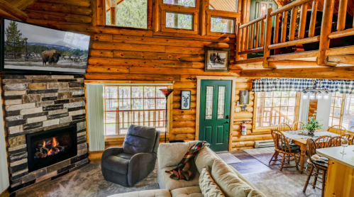 Cozy log cabin interior with a stone fireplace, large windows, and a dining area, featuring rustic wooden decor.